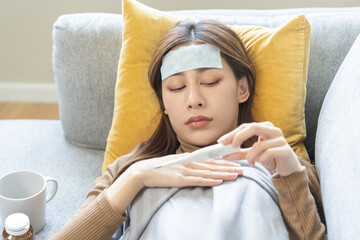 Sick, influenza asian young woman put fever cooling patch on forehead, lying rest on sofa covered body with blanket at home after reading temperature on thermometer, take medicine. Health care person - obrazy, fototapety, plakaty