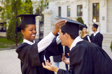 Graduates preparing for graduation. Joyful female student fixing mortarboard to her male classmate...