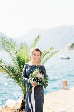 Bride with a bouquet of flowers stands on a pier by the sea against the backdrop of mountains