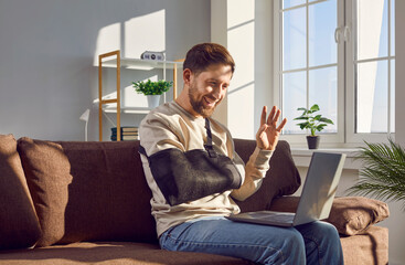 Smiling young man with broken arm making video call using laptop. Cheerful handsome man wearing arm...