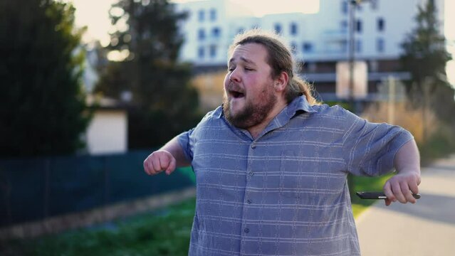 Happy man celebrating success holding phone and walking outdoors dancing in excitement outside in sunlight. One overweight guy feeling excitement