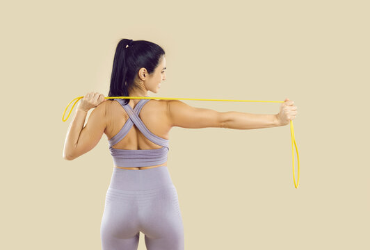 Happy attractive woman in modern sportswear enjoying regular fitness workout, training her body and doing physical exercises with elastic band. Backside view from behind isolated on beige background