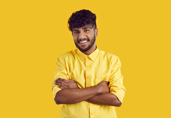 Successful young ethnic Indian man in stylish shirt smiling broadly posing with arms crossed over chest and looking at camera, wearing bright shirt stands in studio with yellow background