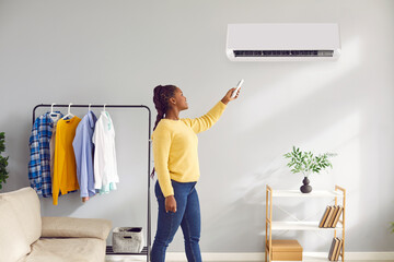Plakat Young woman enjoys a modern air conditioning system at home. Happy beautiful African American woman adjusts temperature on a modern white AC in the living room with a sofa, shelf and clothes rack