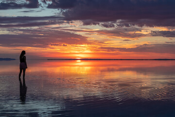 Fototapeta premium Silhouette of woman walking into the sunrise of lake Bonneville Salt Flats, Wendover, Western Utah, USA, America. Dreamy red colored clouds mirroring on the water surface creating romantic atmosphere