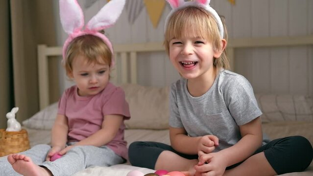 Happy children, baby girl and boy, wearing bunny ears headband playing at home in bedroom with colored Easter eggs. Siblings Enjoying the holidays. Easter day. Christian Passover. Celebrating Easter.