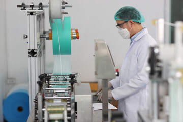 Worker man in personal protective equipment or PPE inspecting quality of mask and medical face mask production line in factory, manufacturing industry and factory concept.