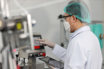 Worker man in personal protective equipment or PPE inspecting quality of mask and medical face mask production line in factory, manufacturing industry and factory concept.