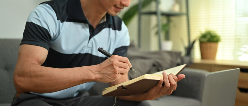 Cropped image of man freelancer sitting on couch and making important notes, planning daily appointment on notepad