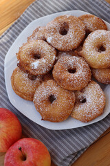 Homemade deep fried apple rings with batter sprinkled with powdered sugar ona white plate on wooden table with fresh apples