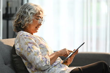 Fototapeta na wymiar Happy elderly gray haired lady enjoy browsing internet on digital tablet, sitting in cozy living room