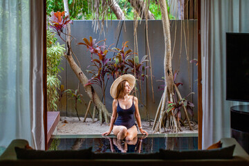 Travel woman in straw hat and black swimsuit sitting on pool edge