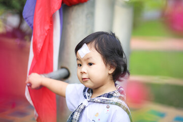 An Asian little girl who wearing a kindergarten uniform and has a wound sewn on her forehead playing alone outside. 