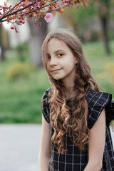 Naklejka premium Young pretty dreaming smiling girl with long hair, sakura branch with gentle pink flowers over her head, looking at camera, warm spring vertical outdoor portrait in park, purity and lightness of youth