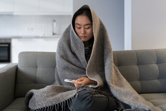 Unhappy Asian Young Female In Warm Plaid Hold Thermometer Suffering From Flu Fever High Temperature On Sofa At Home. Sick Chinese Girl Feeling Unwell Cold Shivering Freezing By Central Heating Problem