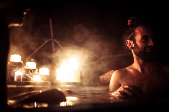 Man with hair in bun sits in out door bath tub in candle light