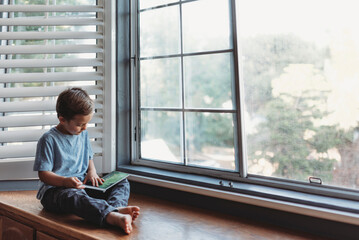 Young boy reads in window alcove with soft light and shutters