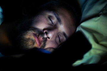 Bearded tired man in bed at night. He is watching something on phone