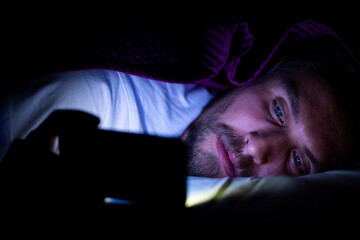 Bearded young man is lying in bed under his blanket looking at phone