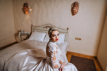A beautiful, young, smiling blonde bride in a white lace long dress sits in a hotel interior room. Wedding photography, portrait.