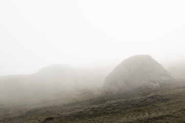 Mountain landscape - silhouette of big grey rounded stones on slope in dense white fog on dim green...