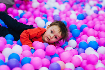 Obraz na płótnie Canvas A beautiful, happy, small, smiling boy, a preschool child lies in a variety of multi-colored, colored plastic balls on the playground. Photography, portrait.