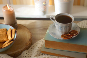 Vintage books, reading glasses, e-reader, cup of tea, bowl of biscuits and lit candle on the table. Selective focus.
