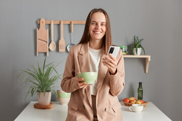 Portrait of charming cute happy young adult woman wearing beige suit standing near table on kitchen at home, eating her dish, using cell phone while having breakfast.