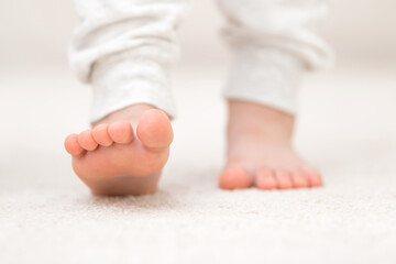 Little child legs in white trousers on light soft carpet background. Barefoot step closeup. Front...