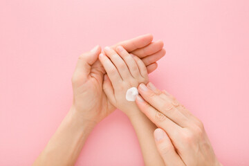 Young adult mother fingers applying white moisturizing cream on little girl hand on light pink...