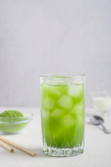 Organic natural antioxidant cold iced matcha cocktail made of green tea leaves powder mixed with water and ice cubes served in drinking glass on white wooden table with ingredients and paper straws