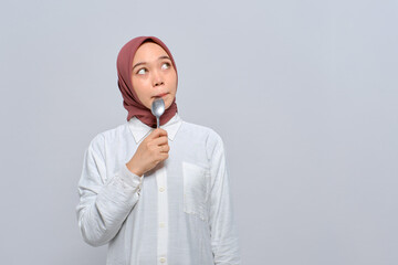Hungry young Asian Muslim woman with spoon in her mouth and looking at copy space, dreaming about delicious healthy food isolated over white background