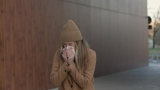 ill beautiful young woman sneezing and blowing nose in napkin. Sick businesswoman in business style sneeze at street. Unhealthy employee. Outdoor. Virus symptoms. Cold disease.