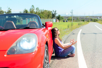 Fototapeta na wymiar Lifestyle portrait of a carefree beautiful woman in sunglasses dressed casually sitting near red cabriolet car on the roadside in relaxation or meditation posture. Road trip enjoying freedom concept 