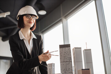 Young female engineer in protective helmet working on building complex prototype project of residential business district measuring city model using compass for drawing at office with panoramic view