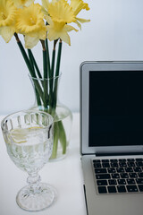 glass of water with lemon, laptop and bouquet of yellow flowers on white desk 