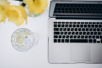 glass of water with lemon, laptop and bouquet of yellow flowers on white desk 