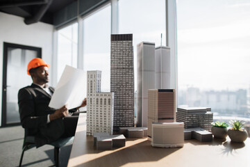 Focus on city buildings model at office interior with panoramic city view, handsome chief architect african man in business suit and protective helmet holding blueprint sitting at desk
