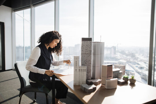 Curly female engineer working on building complex prototype project of residential or business district, measuring blueprint city model using compass at office with panoramic windows