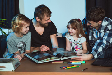 Fototapeta na wymiar Staged photo. Homosexual couple and their children, two cute girls, at home. The whole family is looking intently at the illustrations in the big book. Maybe they're planning a vacation trip.