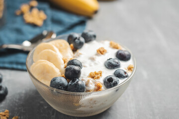 Granola cereal oatmeal with white yogurt, blueberries and banana fruits in a bowl on a blue napkin, grey background