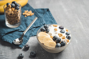 Granola cereal oatmeal with white yogurt, blueberries and banana fruits in a bowl on a blue napkin, grey background