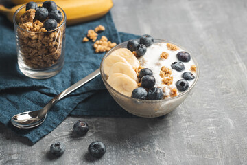 Granola cereal oatmeal with white yogurt, blueberries and banana fruits in a bowl on a blue napkin, grey background