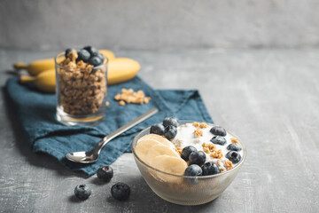 Granola cereal oatmeal with white yogurt, blueberries and banana fruits in a bowl on a blue napkin, grey background