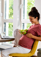Pregnant woman eating salad in front of laptop