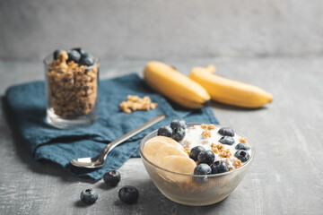 Fototapeta na wymiar Granola cereal oatmeal with white yogurt, blueberries and banana fruits in a bowl on a blue napkin, grey background