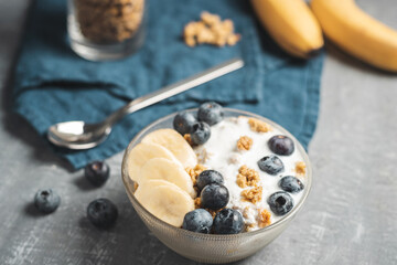 Granola cereal oatmeal with white yogurt, blueberries and banana fruits in a bowl on a blue napkin, grey background