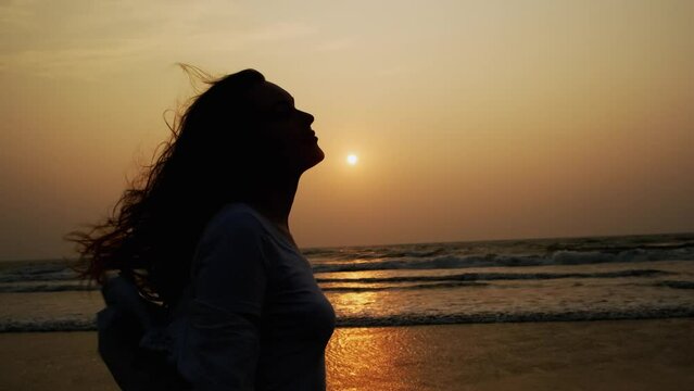 Happy long-haired woman traveller enjoys sea breeze on the seashore at sunset. Female profile silhouette with flowing hair on the setting sun background.