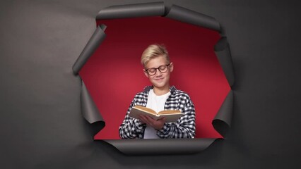 Book lover. Smart teenager boy. Interesting story. Smiling inspired male kid reading literature posing on red background black disrupted paper frame.