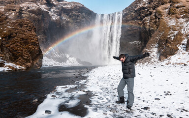 Happy young boy jumping with a snowy waterfall and a rainbow in the background, in Iceland.